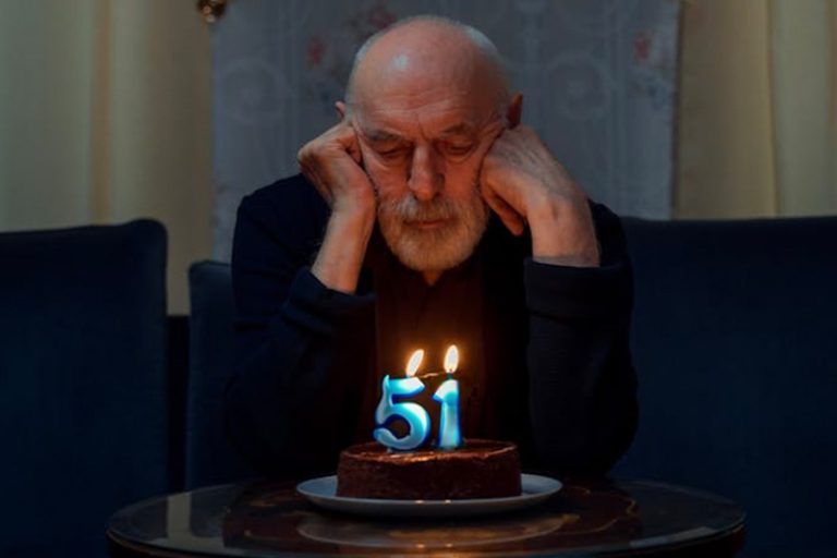 an old man looks at a cake with candles; one candle is a 5 and the other is a 1