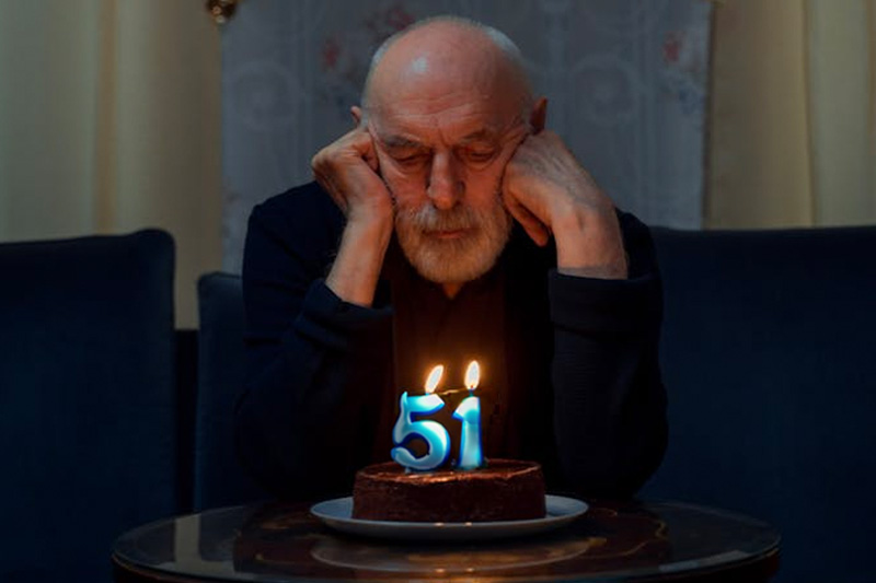 an old man looks at a cake with candles; one candle is a 5 and the other is a 1